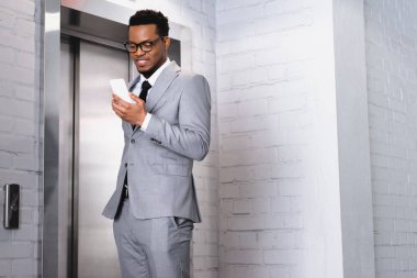 Gülümseyen Afrikalı Amerikalı iş adamı asansörün yanında dururken akıllı telefon tutuyor.