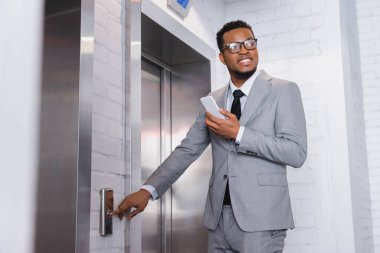 Neşeli Afro-Amerikan iş adamı akıllı telefon tutuyor ve asansör düğmesine basıyor.