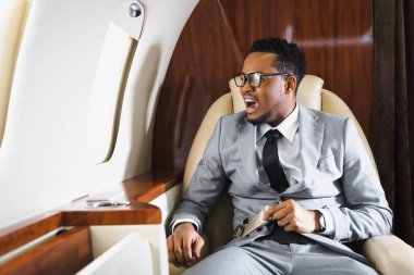 Stresli Afrikalı Amerikalı iş adamı emniyet kemeri tutuyor ve uçuş sırasında panik atak geçiriyor.
