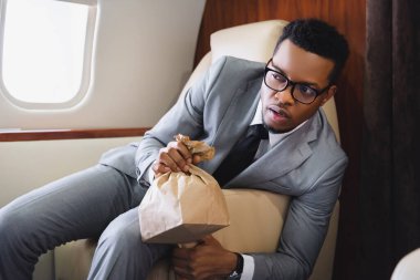 Endişeli Afrikalı Amerikalı iş adamı özel uçakla uçarken panik atak geçirirken kese kağıdı tutuyor.