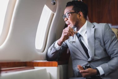 Stresli Afrikalı Amerikalı iş adamı elinde haplar ve bir bardak su tutarken pencereden bakıyor, özel uçakla uçarken panik atak geçiriyor.