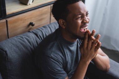 Stresli Afrikalı Amerikalı adam bağırıyor ve evdeki kanepede oturuyor.