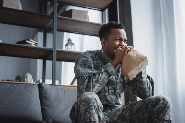 stressed african american soldier in military uniform breathing with paper bag while having panic attack and suffering from PTSD at home