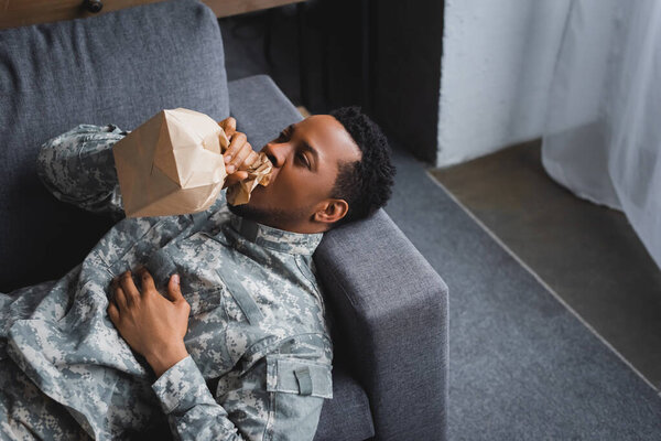 stressed african american soldier in military uniform breathing with paper bag while having panic attack at home
