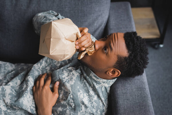 stressed african american soldier in military uniform breathing with paper bag while having panic attack and suffering from PTSD at home