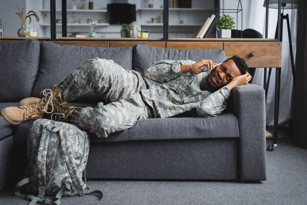 stressed african american soldier in military uniform talking on smartphone and suffering from PTSD at home with backpack