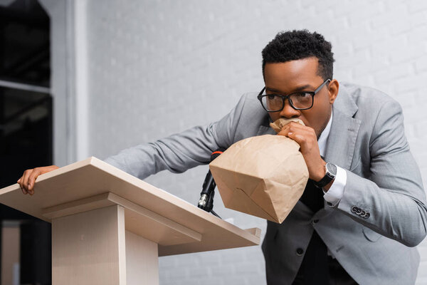 stressed african american speaker breathing with paper bag and having panic attack on business conference 