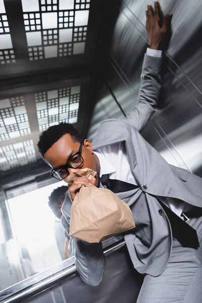 stressed african american businessman breathing with paper bag and suffering from panic attack in elevator