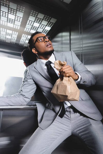 испуганный африканский американский бизнесмен с бумажным пакетом, страдающий от панической атаки в лифте
