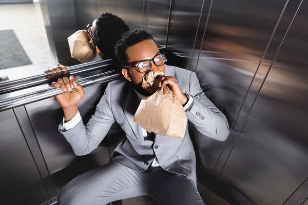 scared african american businessman breathing with paper bag and suffering from panic attack in elevator