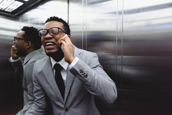 crying african american businessman talking on smartphone and suffering from panic attack in elevator