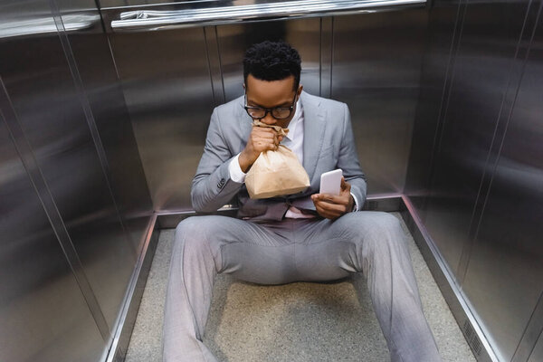 stressed african american businessman with smartphone breathing with paper bag and suffering from panic attack in elevator
