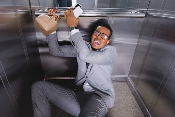 焦虑的非洲裔美国商人 拿着智能手机和纸袋在电梯里遭受恐慌发作 — 图库照片