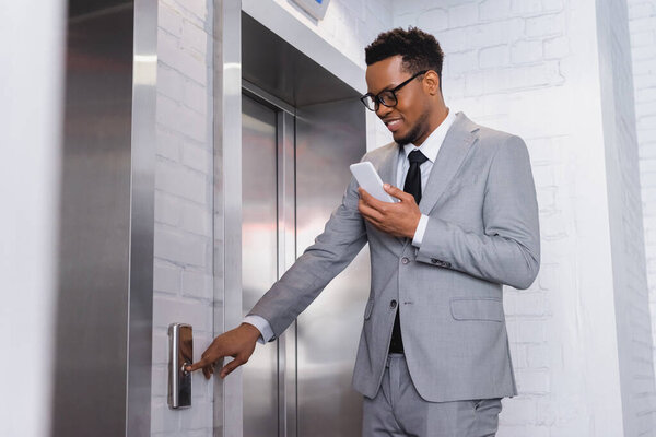 позитивный африканский американский бизнесмен, использующий смартфон во время нажатия кнопки лифта
