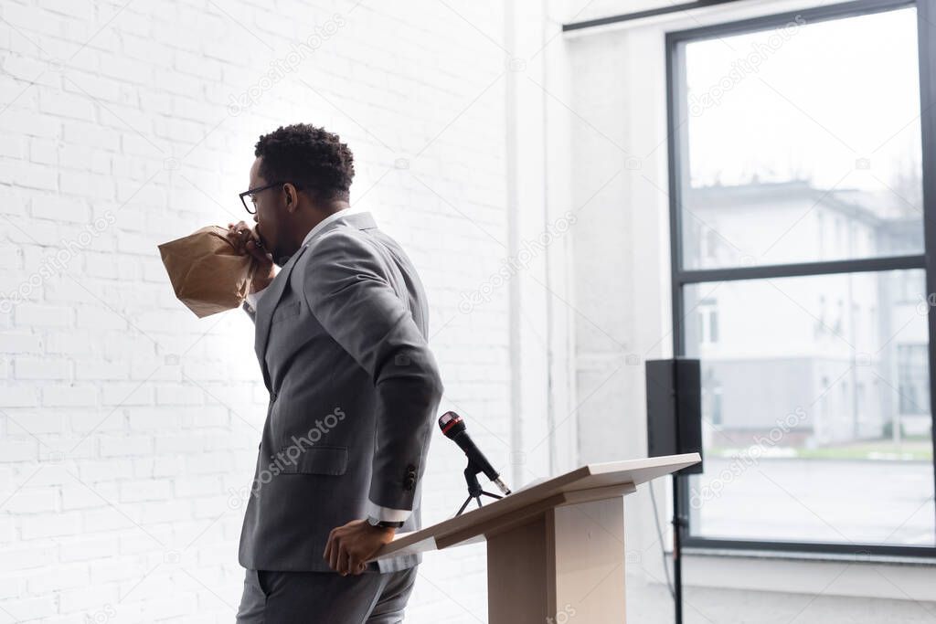 nervous african american speaker breathing with paper bag and having panic attack during business conference in office 
