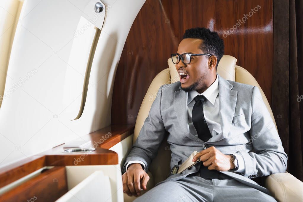 stressed african american businessman holding safety belt and having panic attack during flight on private plane