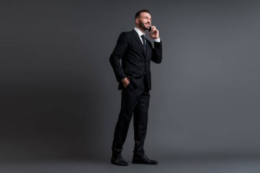 Takım elbiseli mutlu işadamı, gri renkte el ele dururken akıllı telefondan konuşuyor. 
