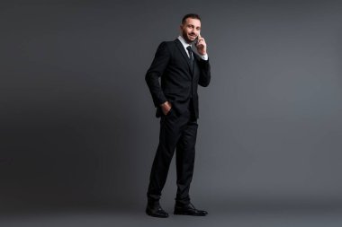 Takım elbiseli neşeli işadamı, gri renkte el ele dururken akıllı telefondan konuşuyor. 