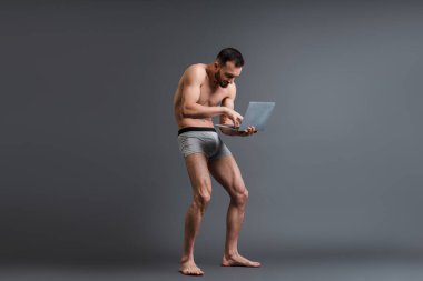 shirtless and stooped man in boxer shorts using laptop on grey  clipart