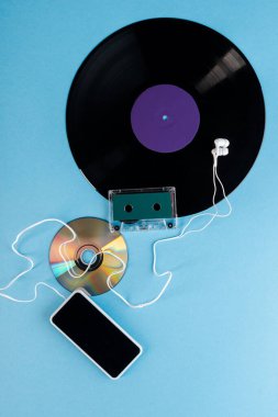 Vinil kaydın üst görüntüsü, CD, kulaklık, ses bandı ve mavi üzerine boş ekran olan akıllı telefon, evrim konsepti 