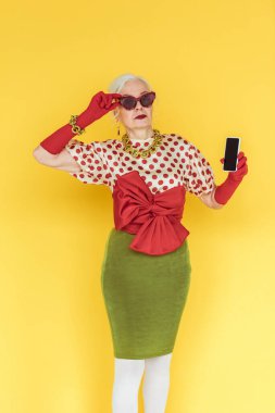 Güneş gözlüklü şık bir son sınıf öğrencisi sarı arka planda akıllı telefon tutuyor.