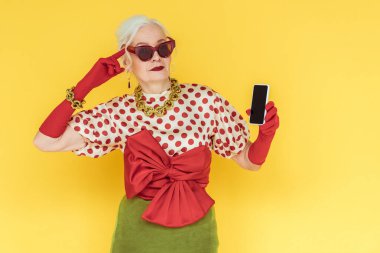 Şık, son sınıf öğrencisi, parmağıyla başını gösteren ve sarıda izole bir akıllı telefon tutan bir kadın.
