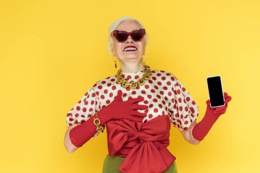 Güneş gözlüklü son sınıf öğrencisi, gülümseyen ve sarıda izole bir akıllı telefon tutan bir kadın.