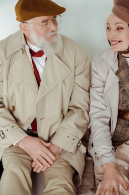 Gri arka planda beyaz bir zeminde otururken birbirlerine gülümseyen son sınıf çifti.