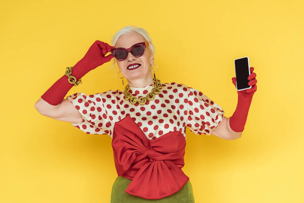 Stylish senior woman smiling and holding smartphone with blank screen on yellow background