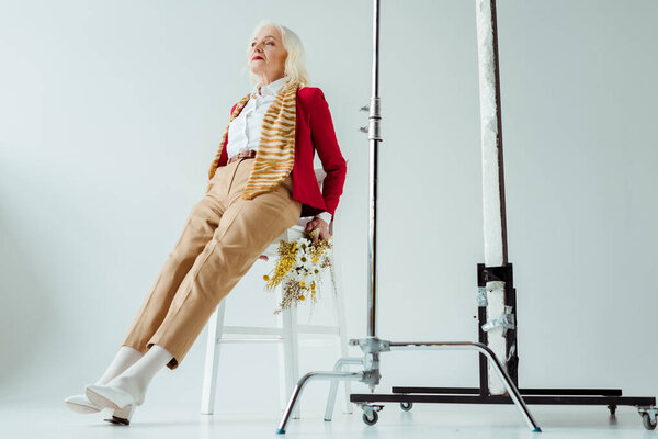 Стильная пожилая женщина с полевыми цветами, сидящая на стуле возле штативов на белом фоне
