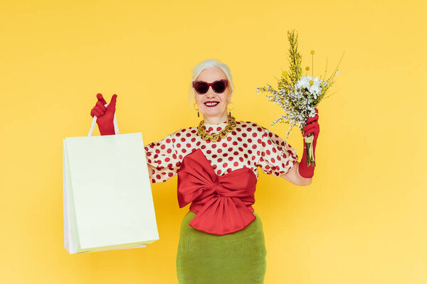 Stylish senior woman smiling while holding wildflowers and shopping bags on yellow background