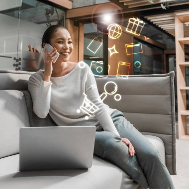 Zeki telefonlu neşeli Asyalı iş kadını dizüstü bilgisayarın yanındaki koltukta otururken konuşuyor.