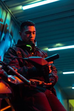 Motosiklet yakınındaki karışık ırk siber punk oyuncusunun seçici odak noktası silaha bakıyor