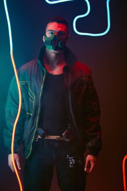 mixed race cyberpunk player in protective mask standing near neon lighting on black  clipart