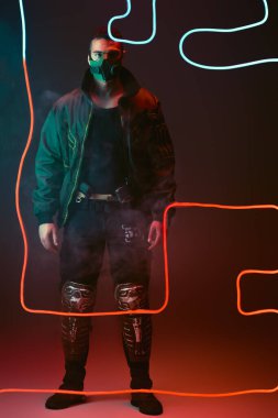 Koruyucu maskeli karışık ırk siber punk oyuncusu siyahın üzerinde kırmızı neon ışığın yanında duruyor. 