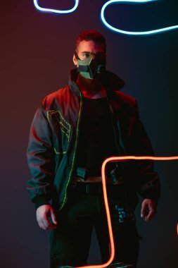 armed and bi-racial cyberpunk player in protective mask standing near neon lighting on black  clipart