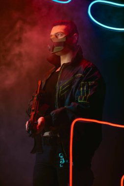 Koruyucu maskeli iki ırklı siber punk oyuncusunun elinde siyah ışıkta dumanlı bir silah var. 