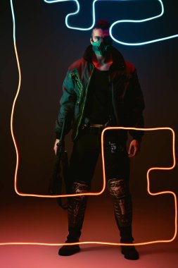 Siyah ışığın yanında dururken koruyucu maskeli karışık ırk siber punk oyuncusu silah tutuyor. 