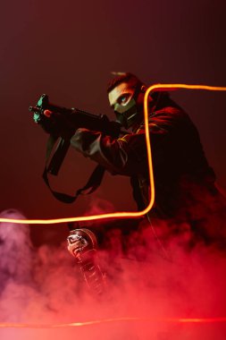 Koruyucu maskeli tehlikeli iki ırklı siber punk oyuncusu siyaha yakın siyaha dumanlı silah doğrultuyor. 