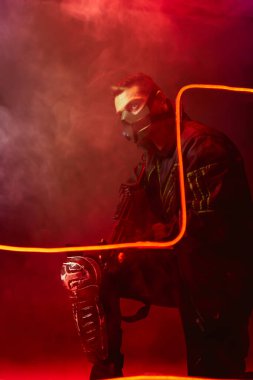 Koruyucu maskeli tehlikeli bir çift ırklı siber punk oyuncusunun elinde siyah ışıkta dumanlı bir silah var. 