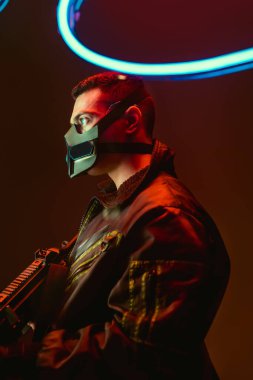 bi-racial cyberpunk player in mask holding gun near blue neon lighting on black  clipart