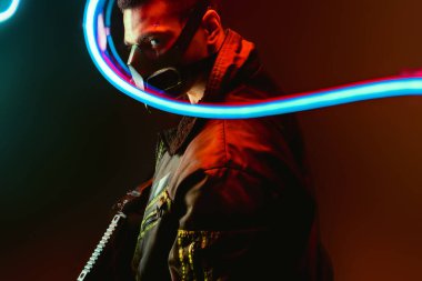 Maskeli tehlikeli karışık ırk siber punk oyuncusunun seçici odak noktası. Elinde mavi neon ışıklı siyah bir silah var. 