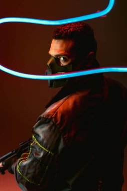selective focus of bi-racial cyberpunk player in protective mask holding gun near blue neon lighting on black  clipart