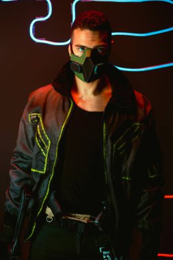 Koruyucu maskeli iki ırklı siber punk oyuncusunun elinde mavi neon ışıklı bir silah var. 