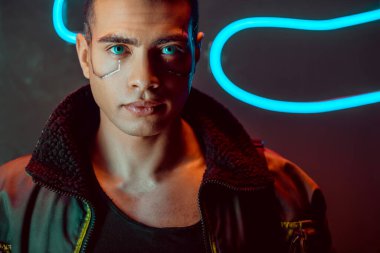 Yakışıklı ve çift ırklı siber punk adam siyah ışıkta, neon ışığın yanındaki kameraya bakıyor. 