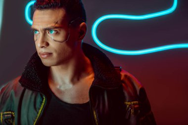 handsome and bi-racial cyberpunk player looking away near neon lighting on black  clipart