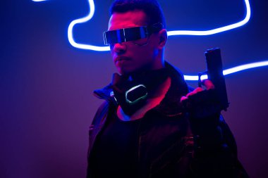 Gelecekçi gözlüklü karışık ırk siber punk oyuncusu mavi neon ışığın yanında silah tutuyor. 