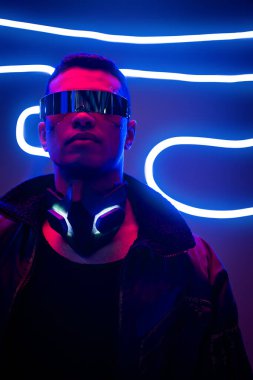 Fütürist gözlüklü karışık ırk siber punk oyuncusu neon ışıklarının yanında duruyor. 