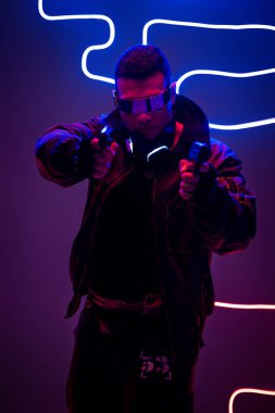 Fütürist gözlüklü yakışıklı melez siber punk oyuncusu neon ışıklarının yanında silah tutuyor. 