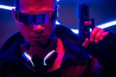 Gelecekçi gözlüklü yakışıklı melez siber punk oyuncusu mavi neon ışığın yanında silah tutuyor. 